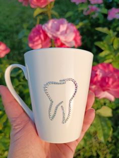 a hand holding a white coffee cup with a tooth design on the side and some pink flowers in the background