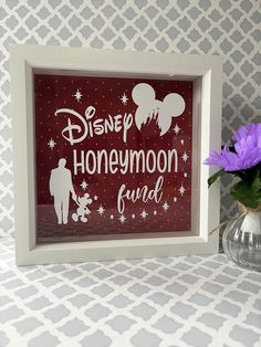 there is a vase with purple flowers in it next to a framed sign that says disney honeymoon fund