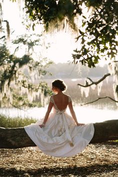 a woman in a white dress sitting on a tree branch looking out over the water