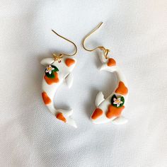 two orange and white cat shaped earrings sitting on top of a white cloth covered surface