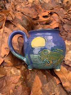 a blue mug with a frog painted on it sitting in the leaves next to a tree