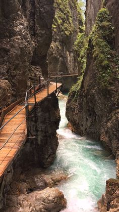 a wooden bridge over a river surrounded by rocky mountains and trees in the middle of it