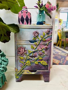 two vases with flowers are on top of a small dresser next to some plants