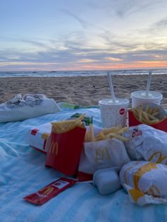 hotdogs, fries and drinks are on the blanket at the beach as the sun sets