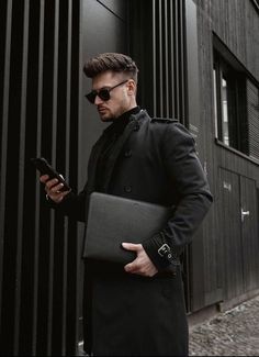 a man in a black coat is holding a laptop and looking at his cell phone