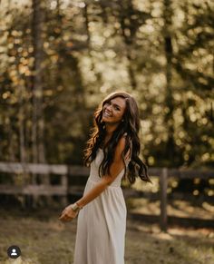 a woman in a white dress smiles as she walks through the woods with her hands behind her back