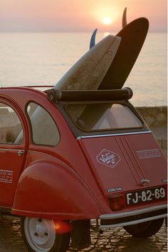 a small red car with a surfboard on the roof parked in front of the ocean