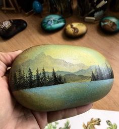 a hand holding a painted rock with mountains and pine trees on it in front of other rocks