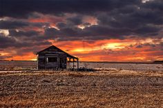 an old shack sitting in the middle of a field under a cloudy sky at sunset