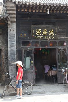 a man standing in front of a restaurant with a bicycle parked outside the entrance and people sitting at tables