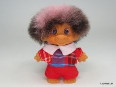 a small doll with pink hair wearing a red shirt and blue plaid pants, standing on a white surface