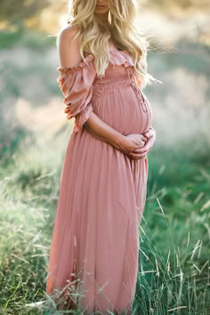 a pregnant woman wearing a pink dress standing in tall grass with her hands on her stomach