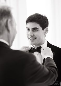 a man in a tuxedo adjusts his bow tie while another man looks on