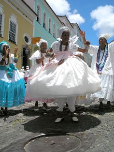 three women in white dresses are dancing on the street with other people dressed in blue and white