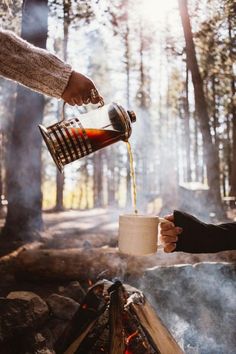 someone is pouring something into a cup over a campfire in the woods while another person holds out their hand