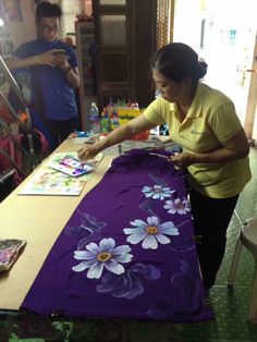 a woman in yellow shirt painting on purple table cloth with white daisies and blue flowers