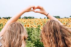 two women making heart shape with their hands in front of a field of sunflowers