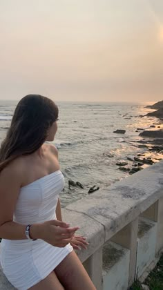 a woman in white dress sitting on the edge of a wall next to the ocean