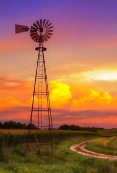 a windmill sitting on top of a lush green field next to a dirt road in front of a sunset