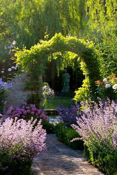 a garden with lots of flowers and trees in the background, including an arch over a stone path