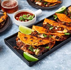 some tacos are on a black plate with lime wedges and salsa in the background