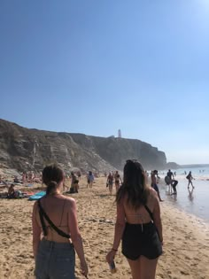 two women in bathing suits walking on the beach towards the ocean with people standing around