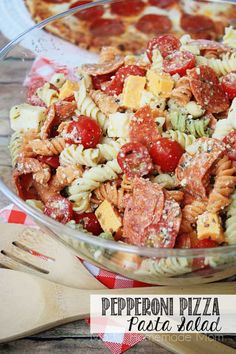 a pasta salad with pepperoni and tomatoes in a glass bowl next to a pizza