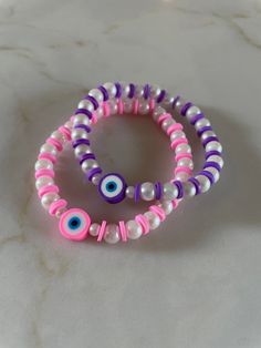 two bracelets with evil eyes and pink, purple, and white beads on a marble surface