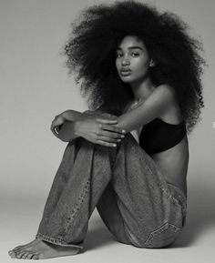 a woman with an afro sitting on the ground wearing jeans and a tank top, looking off to the side