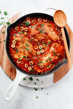 a skillet filled with chicken and olives on top of a wooden cutting board