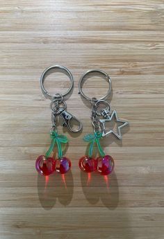 two cherry shaped key chains with charms attached to them on a wooden surface, one has a star and the other has an arrow