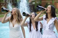 three women in white dresses are drinking from bottles near a fountain with water spouting behind them