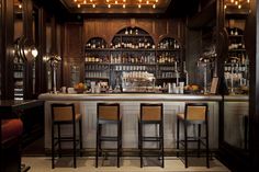 a bar with several stools and bottles on the shelves in front of it is lit by lights