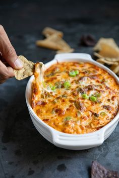 a hand holding a tortilla chip and dipping it into a casserole dish