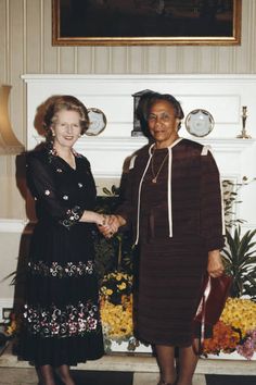 two women standing next to each other in front of a fireplace with flowers on the mantle