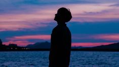 a man standing in front of a body of water at dusk with the sun setting