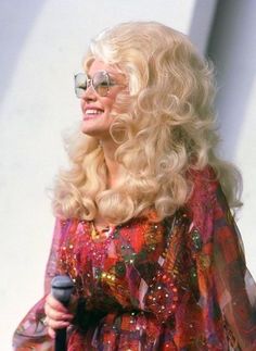 a woman with long blonde hair and glasses holding a microphone in her right hand while standing next to a white wall