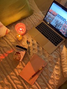 an open laptop computer sitting on top of a bed next to a notebook and candle