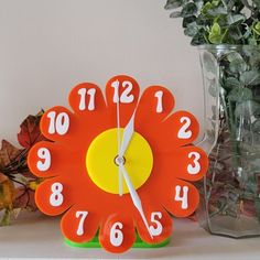 an orange flower shaped clock sitting on top of a white shelf next to a vase with flowers