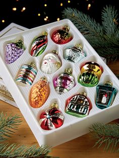 an assortment of christmas ornaments in a white box on a table next to pine branches