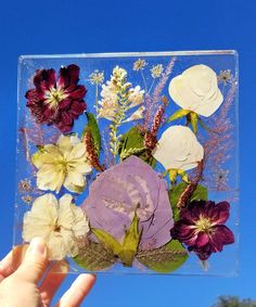 a hand holding up a clear square with flowers in it and leaves on the bottom