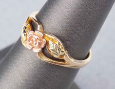 Slim band solid gold ring, featuring an open rose gold bud blossom. Very fine vintage condition with minimal light wear from normal use. Size is 6 US. Feminine ring design showcases Landstrom's & Black Hills Gold's use of 3 colors of solid gold.  A 6mm natural looking open blossom rose flower is in the center of the ring. Edges of the rose petals are highlighted with yellow gold. A 12K green gold leaf on a 12K rose gold leaf sits to each side of the 12K red gold flower. The 2mm narrow yellow gol Hallmarked 14k Gold Flower Ring, Gold 14k Hallmarked Flower Ring, Spring Flower-shaped Gold Rings, Yellow Gold Flower-shaped Jewelry With Rose Design, Black Hills Gold Rings Vintage, Black Hills Gold Rings, Minimal Light, Open Flower, Open Rose