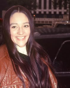 a woman with long brown hair wearing a leather jacket and smiling at the camera in front of a car