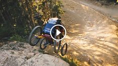 a man riding a bike down a dirt road next to a lush green forest on top of a rock covered hillside