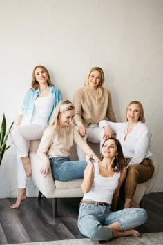 four women are sitting on a white chair in front of a wall and a potted plant