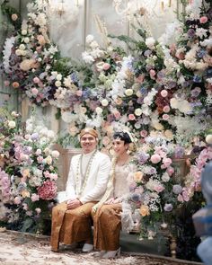 a man and woman sitting on a bench in front of a floral wall with flowers