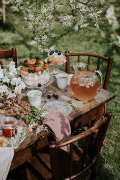 a wooden table topped with plates and bowls filled with fruit next to flowers on top of a lush green field