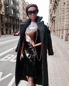 a doll is walking down the street with a handbag on her shoulder and wearing sunglasses