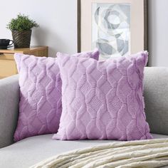 two purple pillows sitting on top of a gray couch next to a white throw pillow