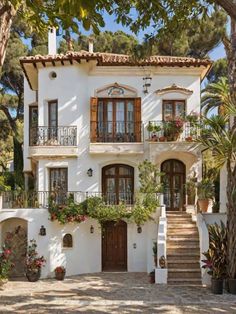 a white house with lots of windows and plants on the front porch, surrounded by trees